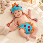 Awake newborn wearing the blue and orange Lumiwee dinosaur crochet photo props beside a parent's hand